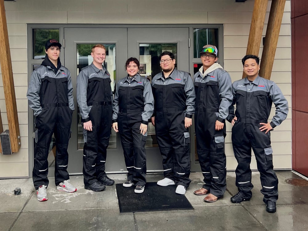 Six people standing in front of a building entrance, wearing matching gray and black coverall uniforms. They are positioned in a row on a wet sidewalk, with double doors and wooden structural beams behind them.