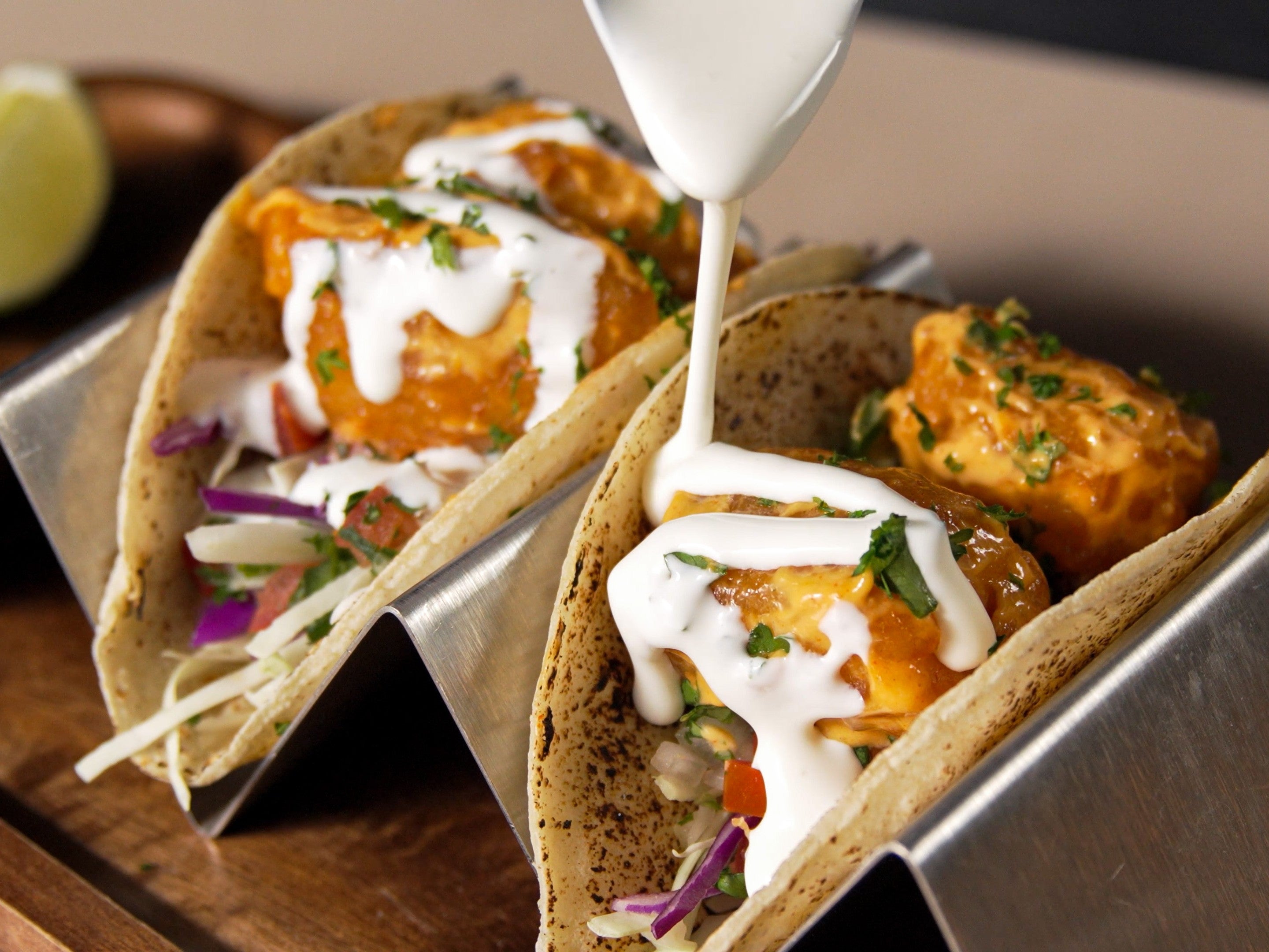 Two tacos in metal holders filled with crispy fish, shredded cabbage, diced vegetables, and fresh herbs, topped with creamy white sauce being drizzled over them, with a lime wedge in the background.