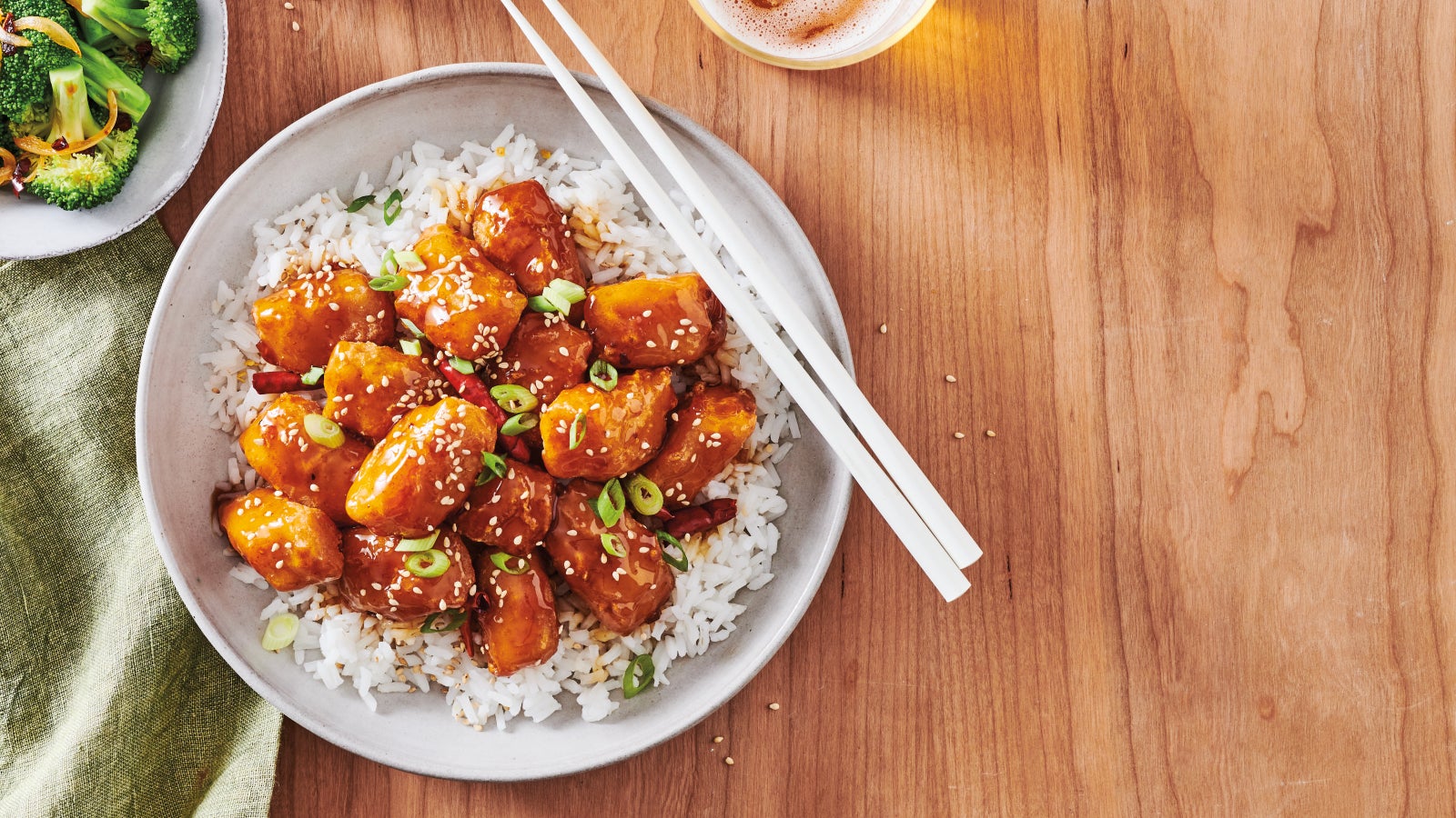 Orange-glazed alaska pollock nuggets served over white rice, topped with sesame seeds and green onions, with chopsticks on the plate. A side of sautéed broccoli, sesame seeds, and a glass of beer on a wooden table.