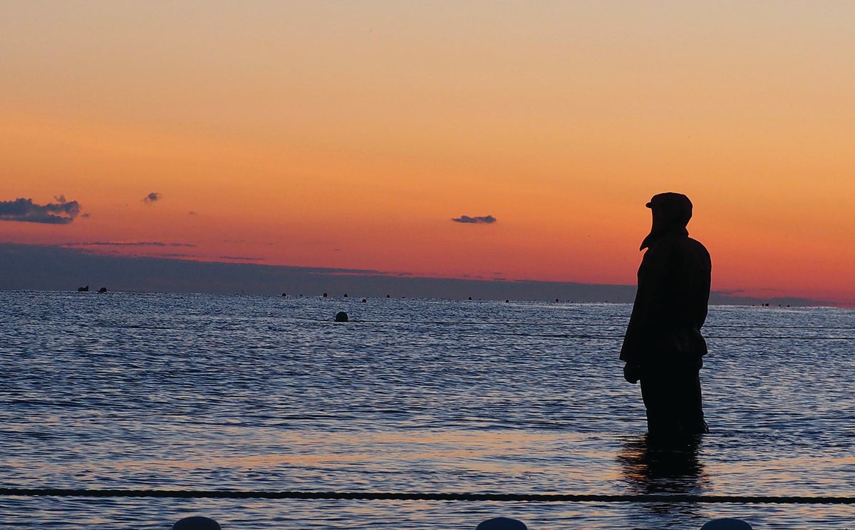 An Alaskan fisherman stands in knee-deep water, looking into the distance at an Alaskan sunrise. 