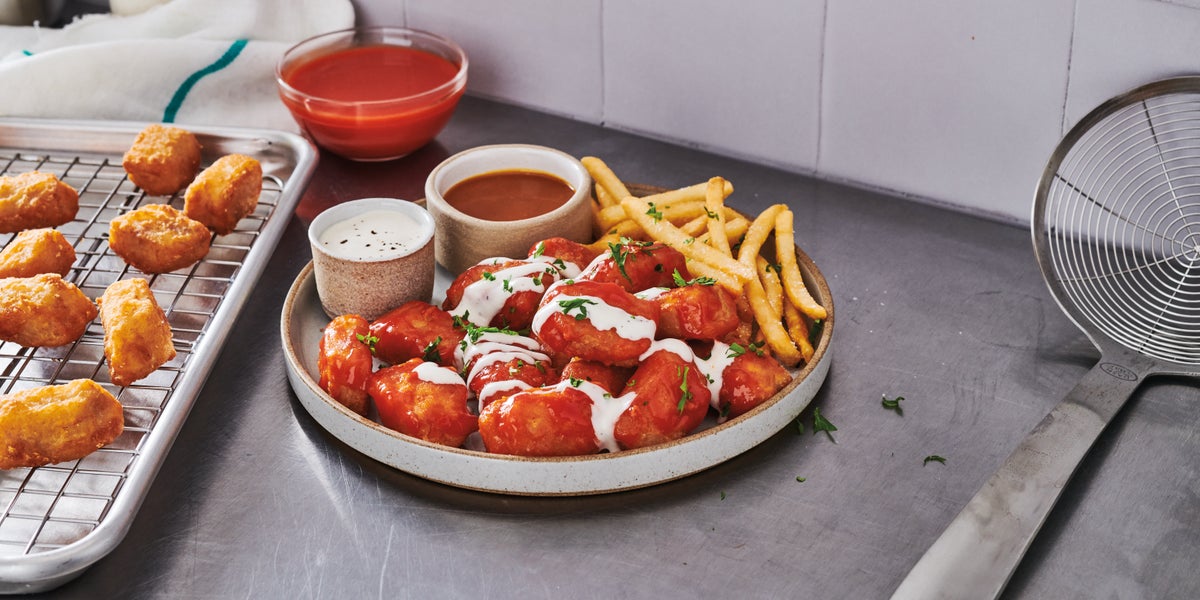 Plate of saucy buffalo-style alaska pollock nuggets drizzled with ranch dressing, served with French fries and two dipping sauces, next to a tray of plain nuggets on a wire rack and a bowl of bright red buffalo sauce on a stainless steel countertop.
