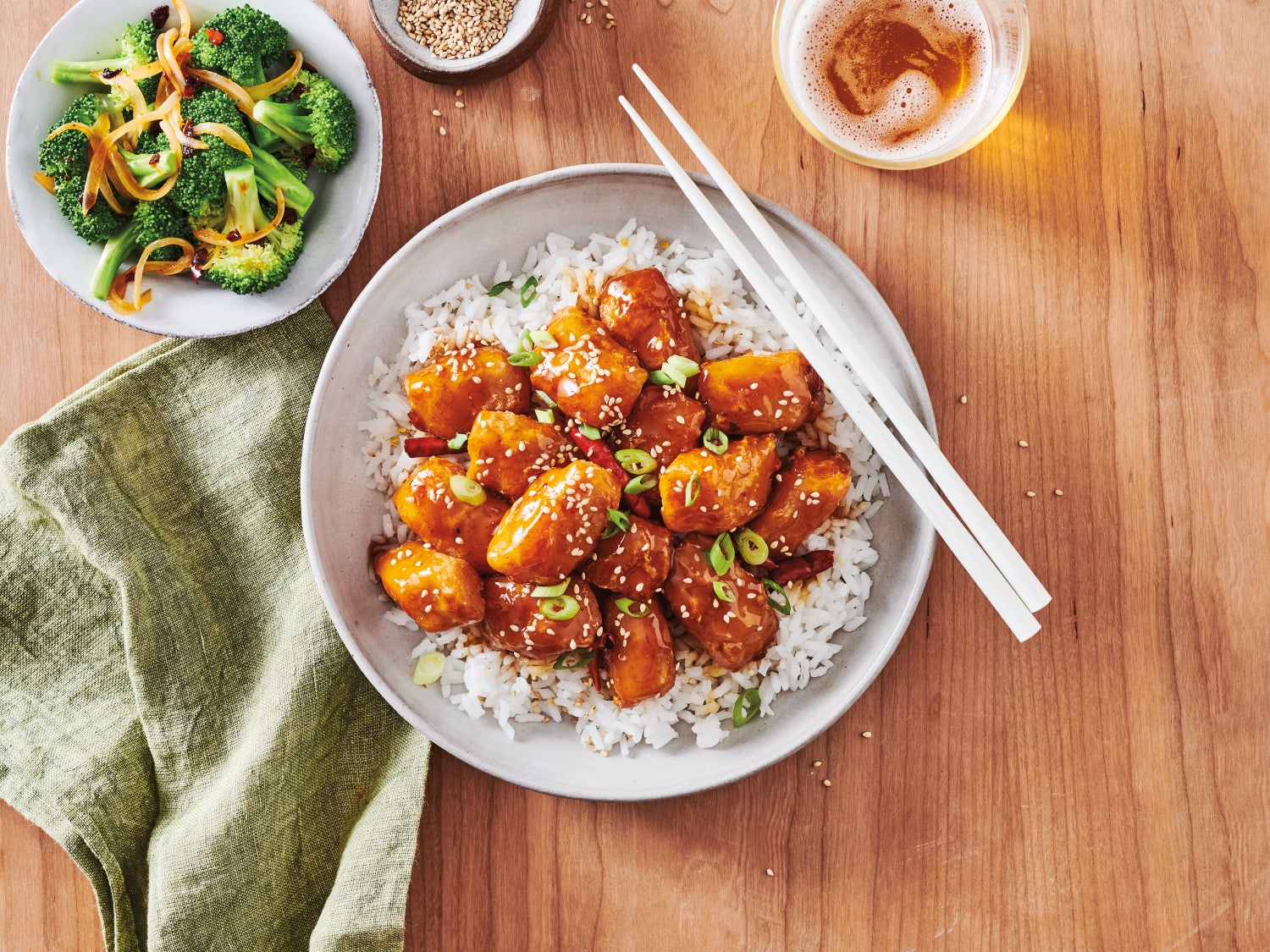 Orange-glazed alaska pollock nuggets served over white rice, topped with sesame seeds and green onions, with chopsticks on the plate. A side of sautéed broccoli, sesame seeds, and a glass of beer on a wooden table.