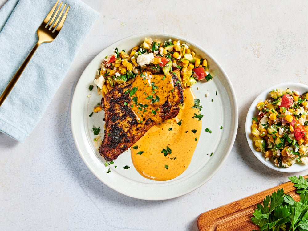 Overhead view of a blackened Alaska cod fillet with creamy orange sauce, paired with a colorful corn salad featuring zucchini, tomatoes, herbs, and crumbled cheese.