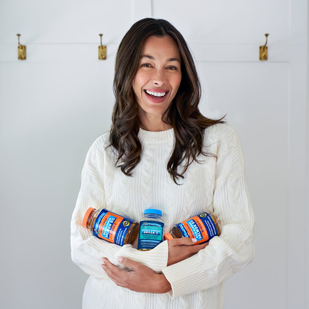 A woman holding three bottles of Pure Alaska Omega cradled in her arms wearing a white sweater with a off white background with coat hooks across the wall.