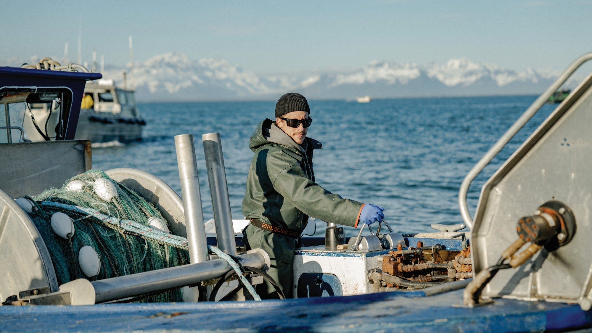 Crew member aboard a commercial fishing boat working near fishing nets and deck machinery, with a calm ocean and snow-covered mountains along the Alaska coastline in the distance.