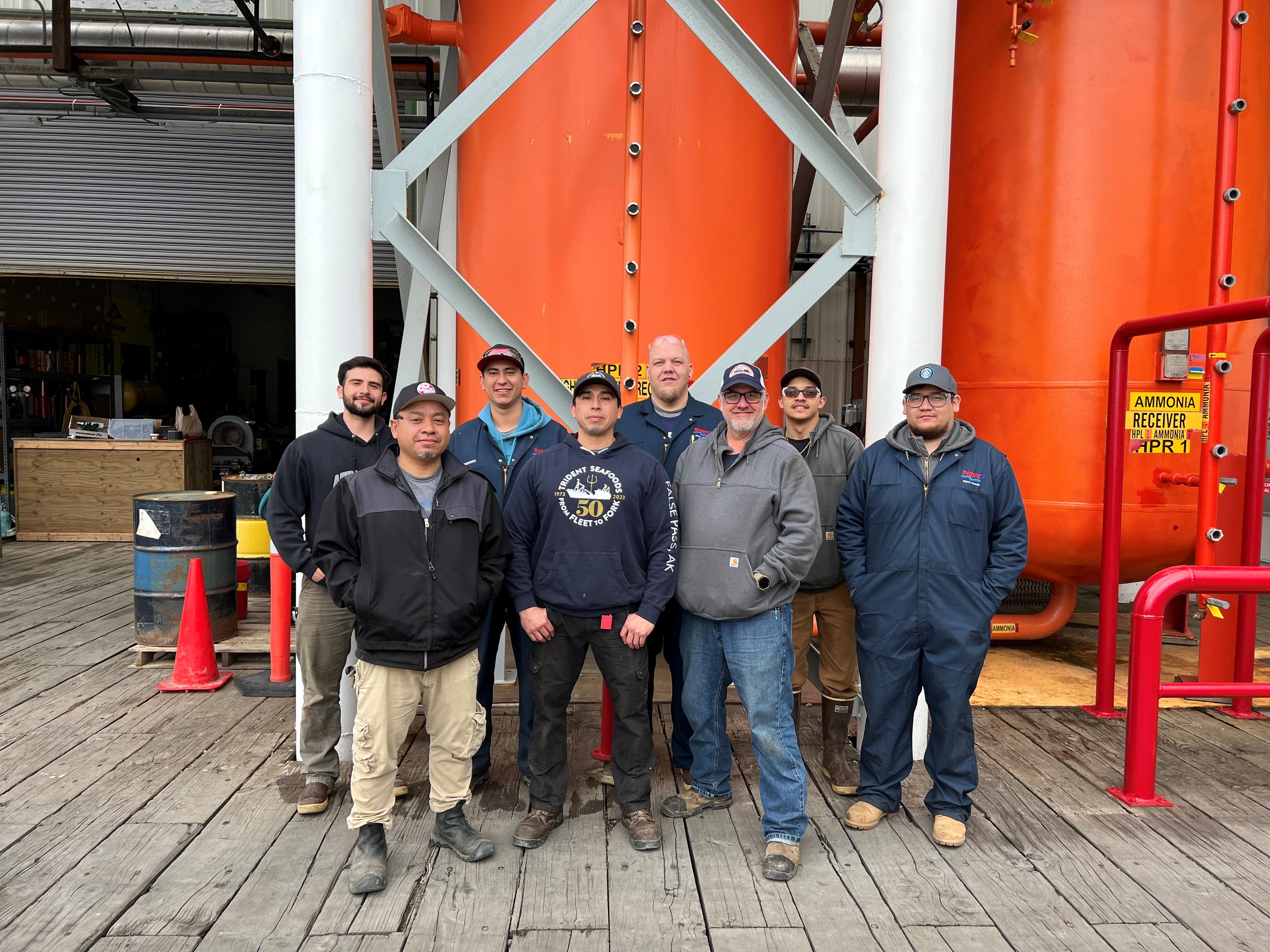 Skilled trades technicians pose in front of orange refrigeration tanks at Trident Seafoods' Naknek, Alaska plant. 