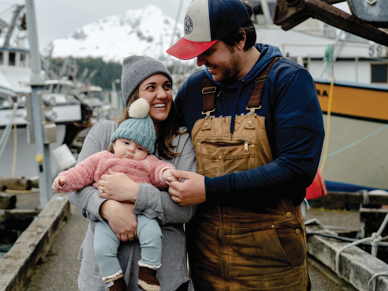 Two adults and an infant dressed in cold-weather clothing stand on a working harbor dock beside fishing vessels, with a coastal Alaska mountain landscape behind them.