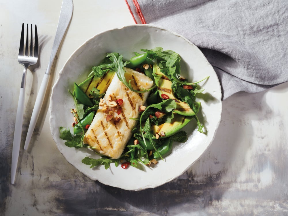 Overhead view of a grilled Alaska cod fillet atop a green salad of arugula, grilled avocado slices, snap peas, fresh herbs, and chopped nuts, served in a white bowl on a light tabletop with a fork, knife, and linen napkin.