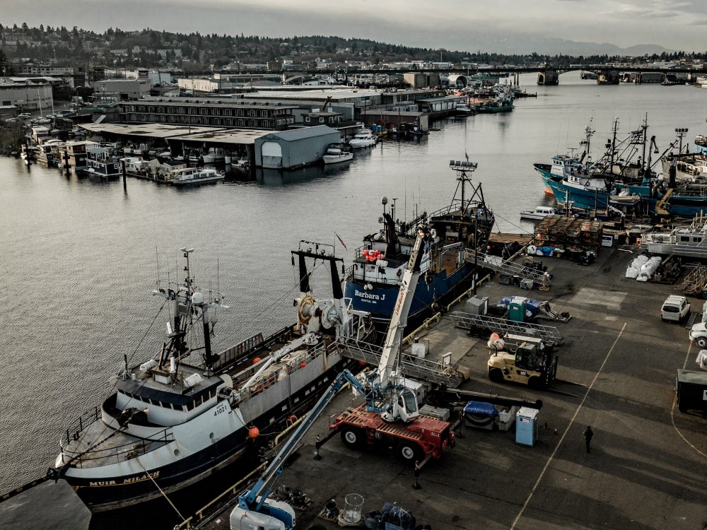 Aerial view of a busy Seattle waterfront industrial harbor with fishing vessels docked along a pier, cranes and equipment on the wharf, warehouses lining the shoreline, and the Ballard bridge spanning the waterway under an overcast sky.