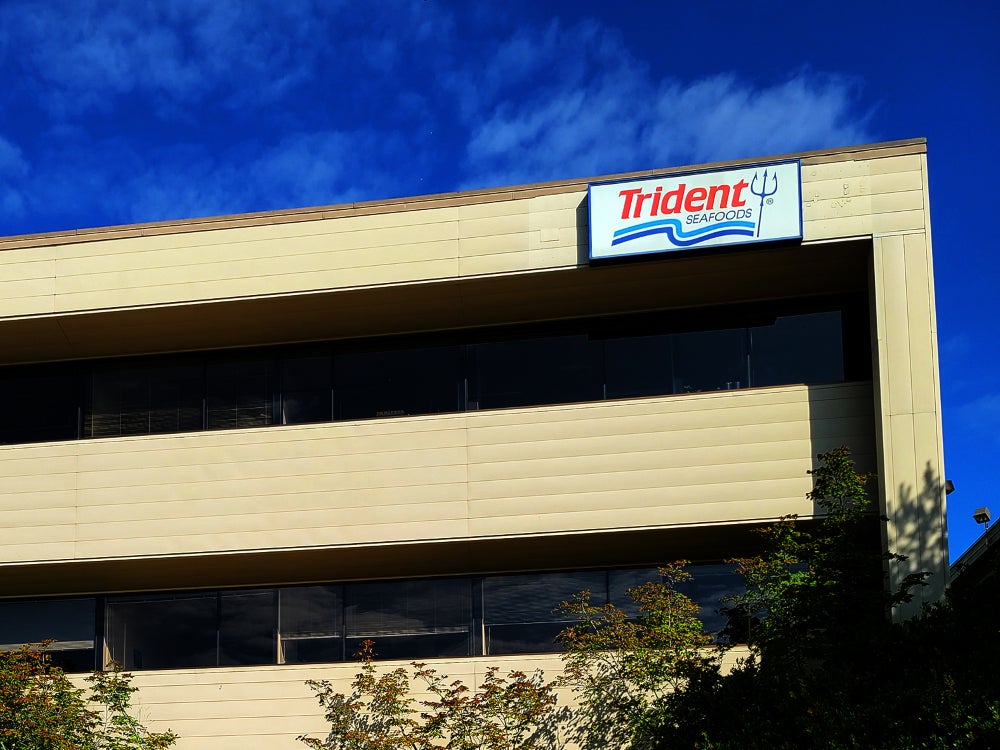 Exterior view of a multi-story office building with a Trident Seafoods sign mounted near the top corner. The building features long horizontal windows, light-colored panels, and landscaping in the foreground beneath a bright blue sky with scattered clouds.