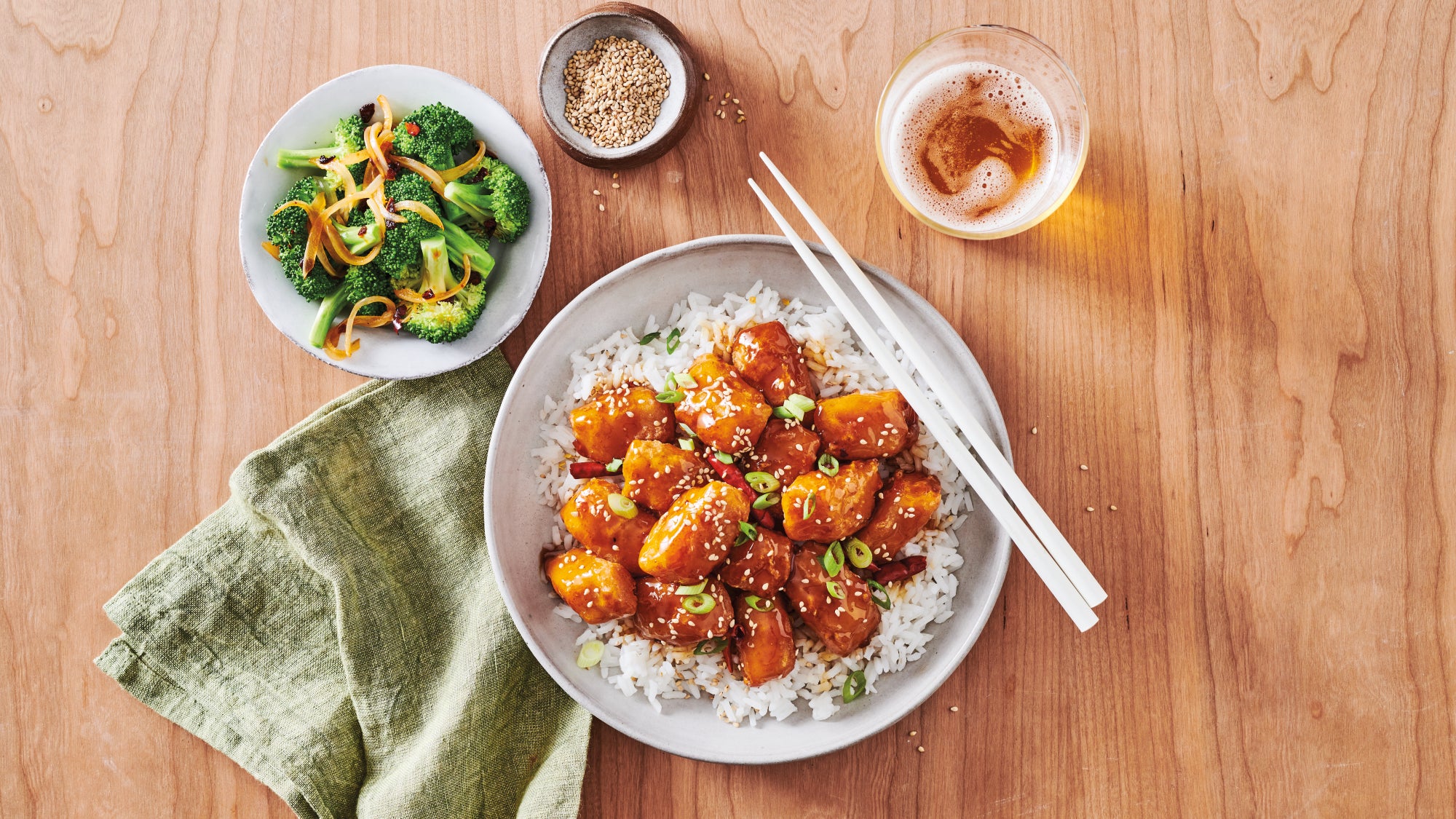 Plate of orange glazed wild Alaska pollock nuggets with sesame seeds and green onions over white rice, with chopsticks on the side. Nearby are a small plate of sautéed broccoli, a bowl of sesame seeds, and a glass of amber drink on a wooden table.