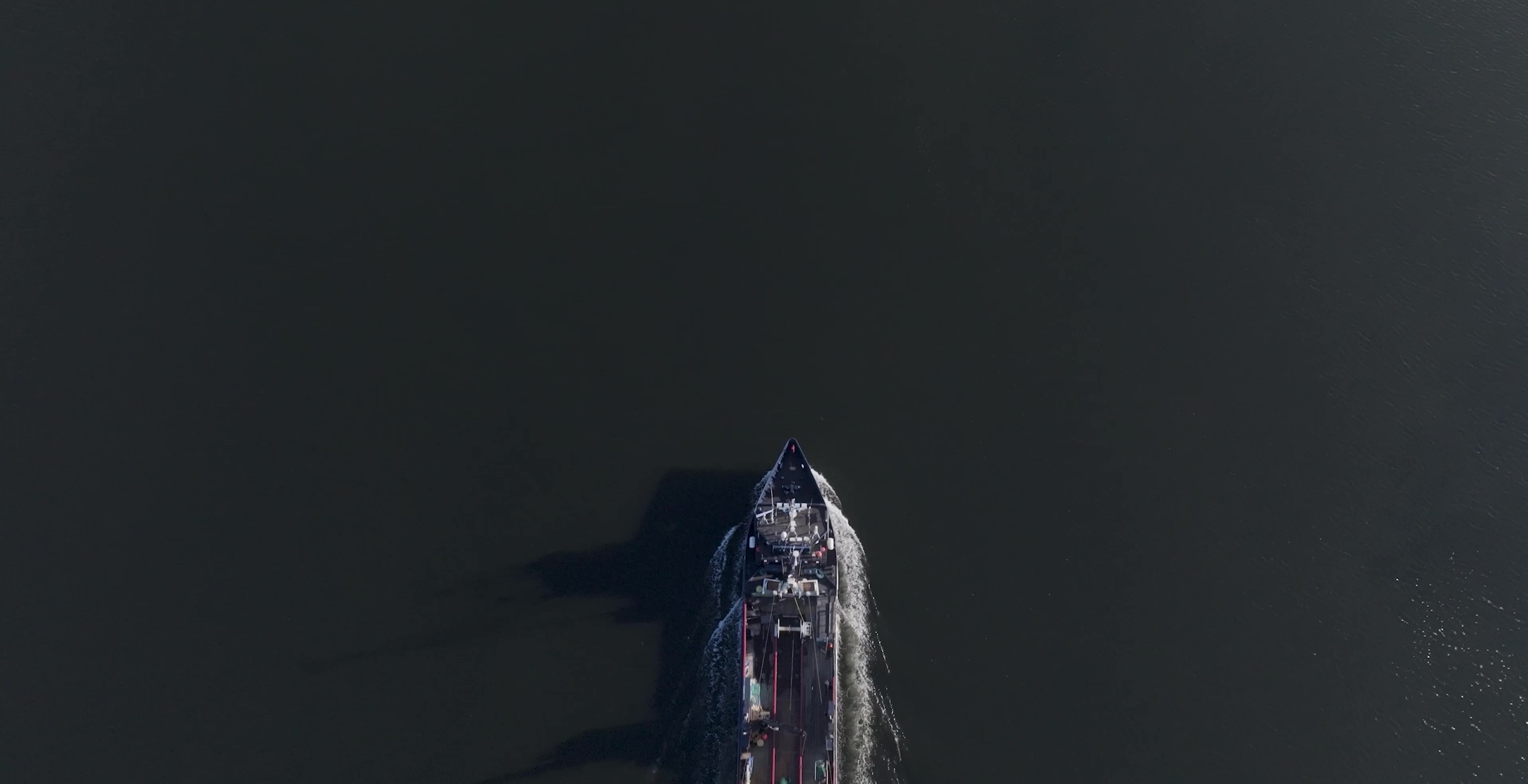 Aerial, top-down view of a large ship moving through dark, calm water, creating a narrow wake, with extensive open water surrounding the vessel.