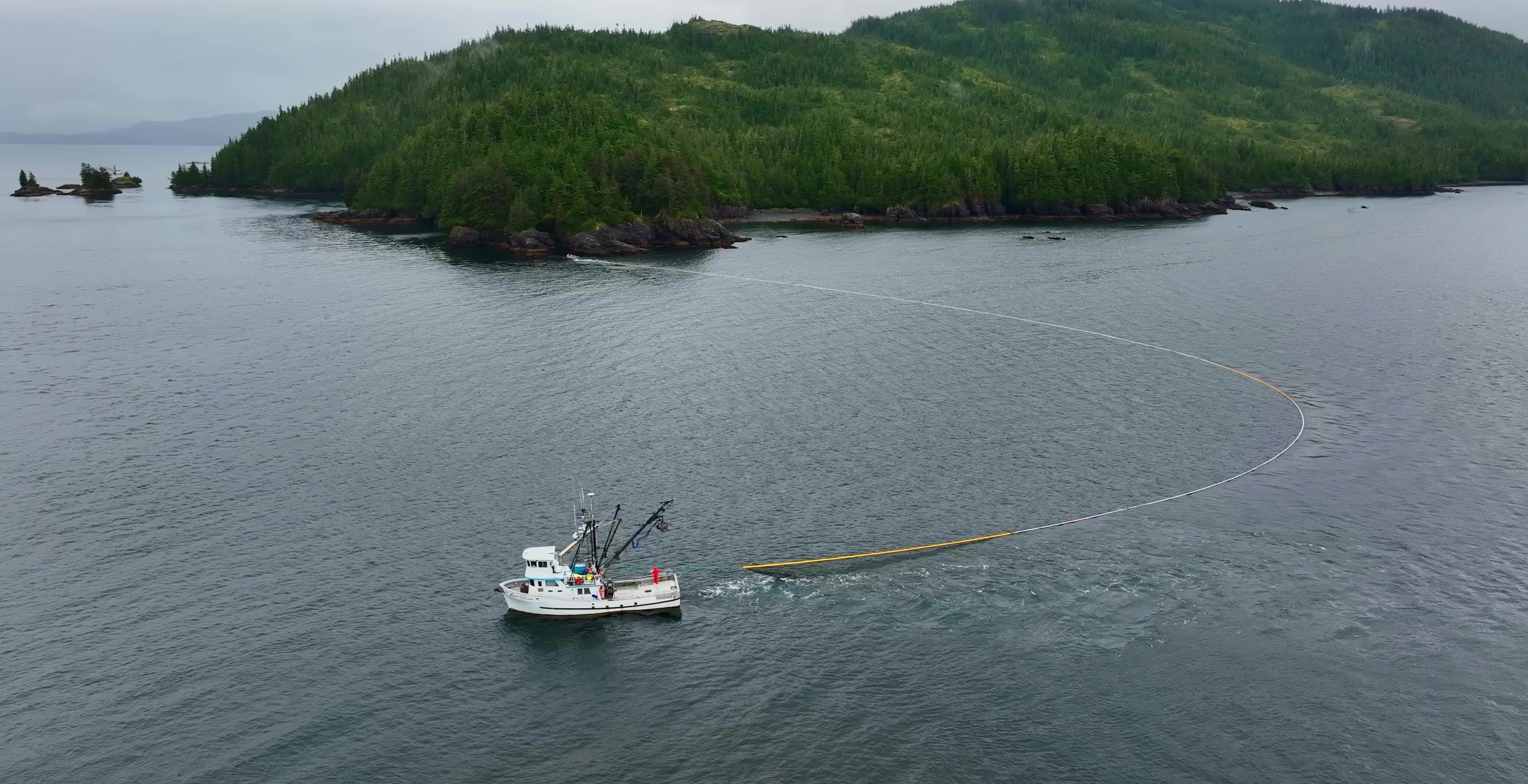 Aerial view of a purse seine vessel deploying a long net in calm coastal waters near a forested island shoreline under overcast skies.