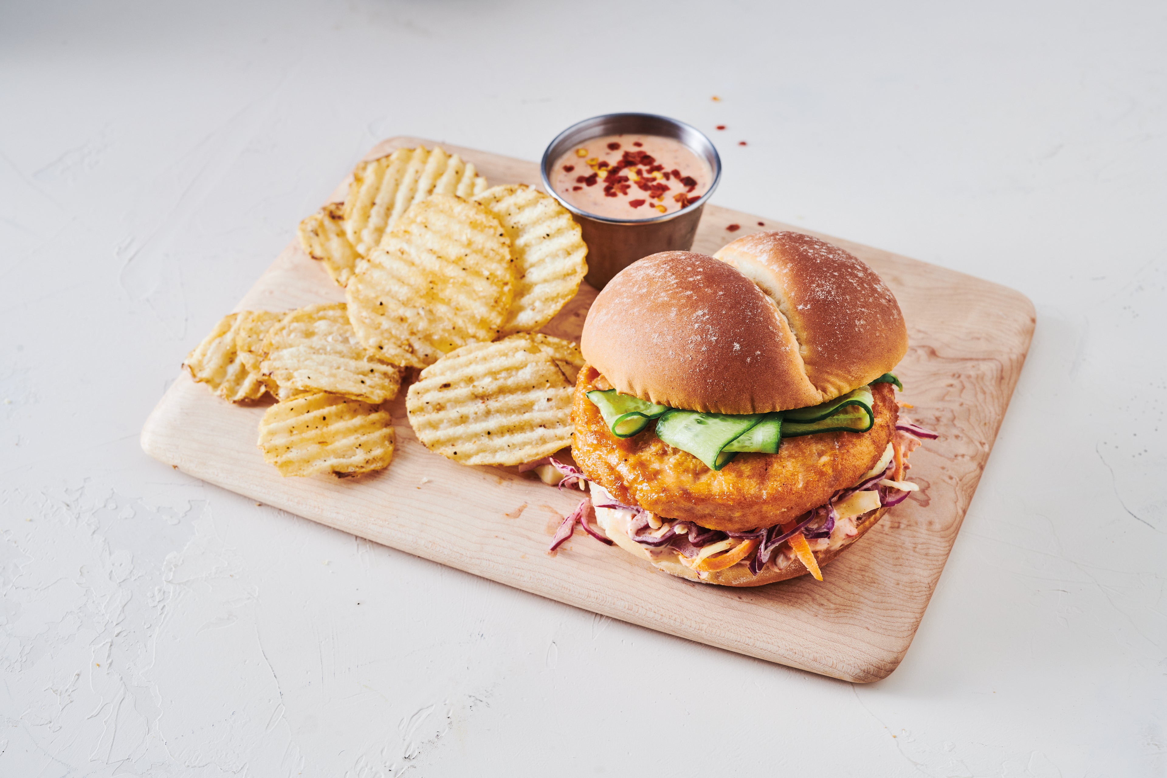 Salmon burger in a bun with various condiments presented on a wooden board with crinkle cut potato chips on the side. 
