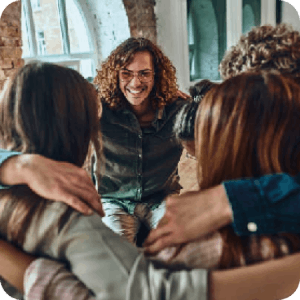 From over the shoulder of a person standing in a circle where everyone has their arms over each other's shoulders, a person is smiling back at the rest of the group.
