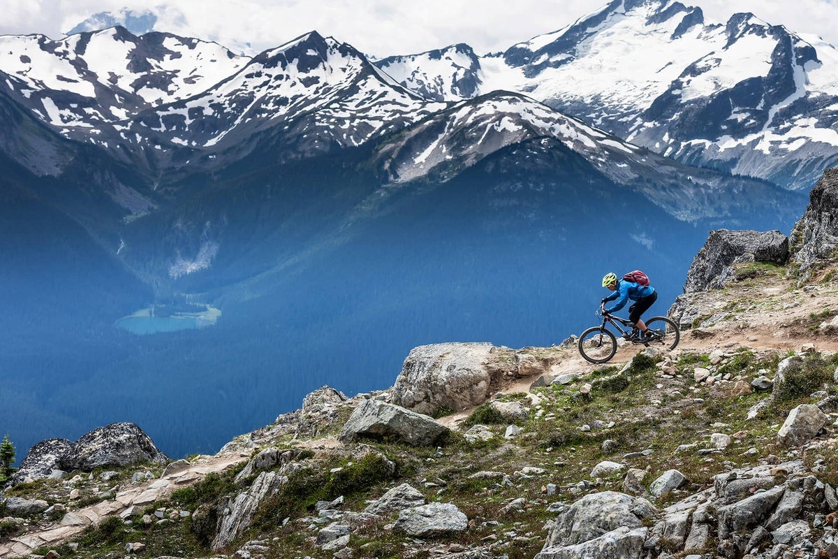 Person biking down a narrow trail alongside rocks with snow-covered mountains behind them