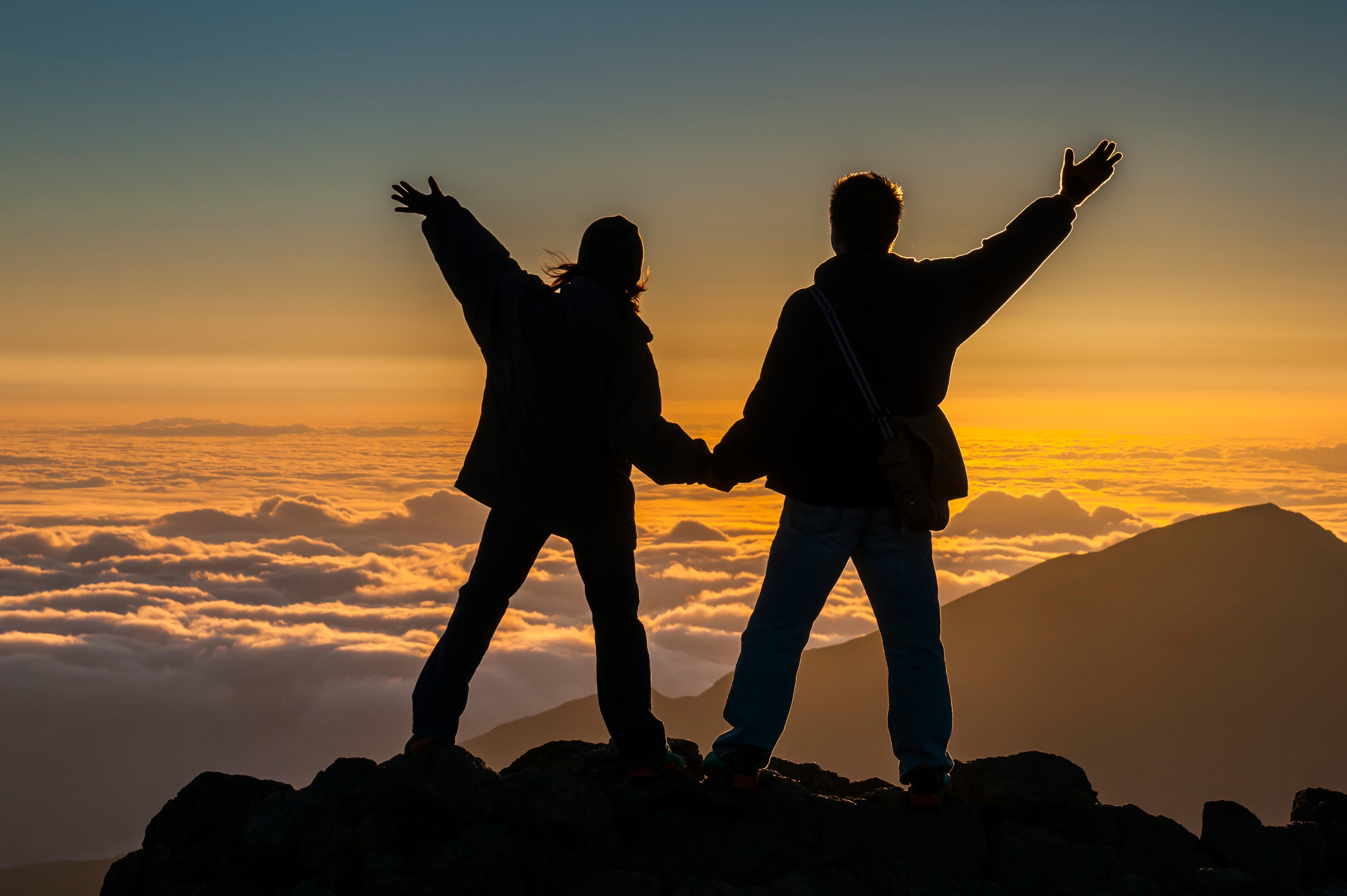 Two people at top of summit overlooking mountain and clouds at sunset