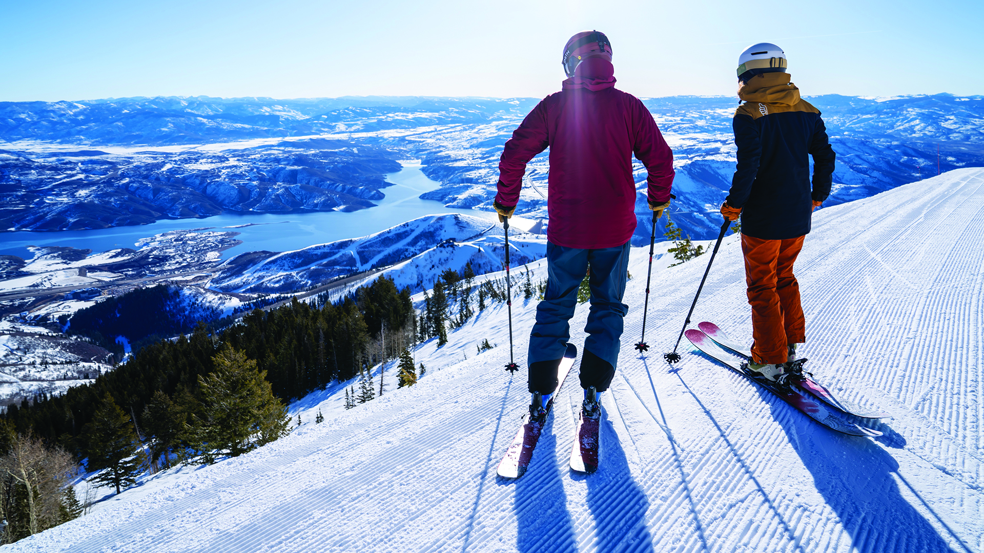 Skiers taking a break on groomed ski run while looking down at lake and snow-covered rolling hills.