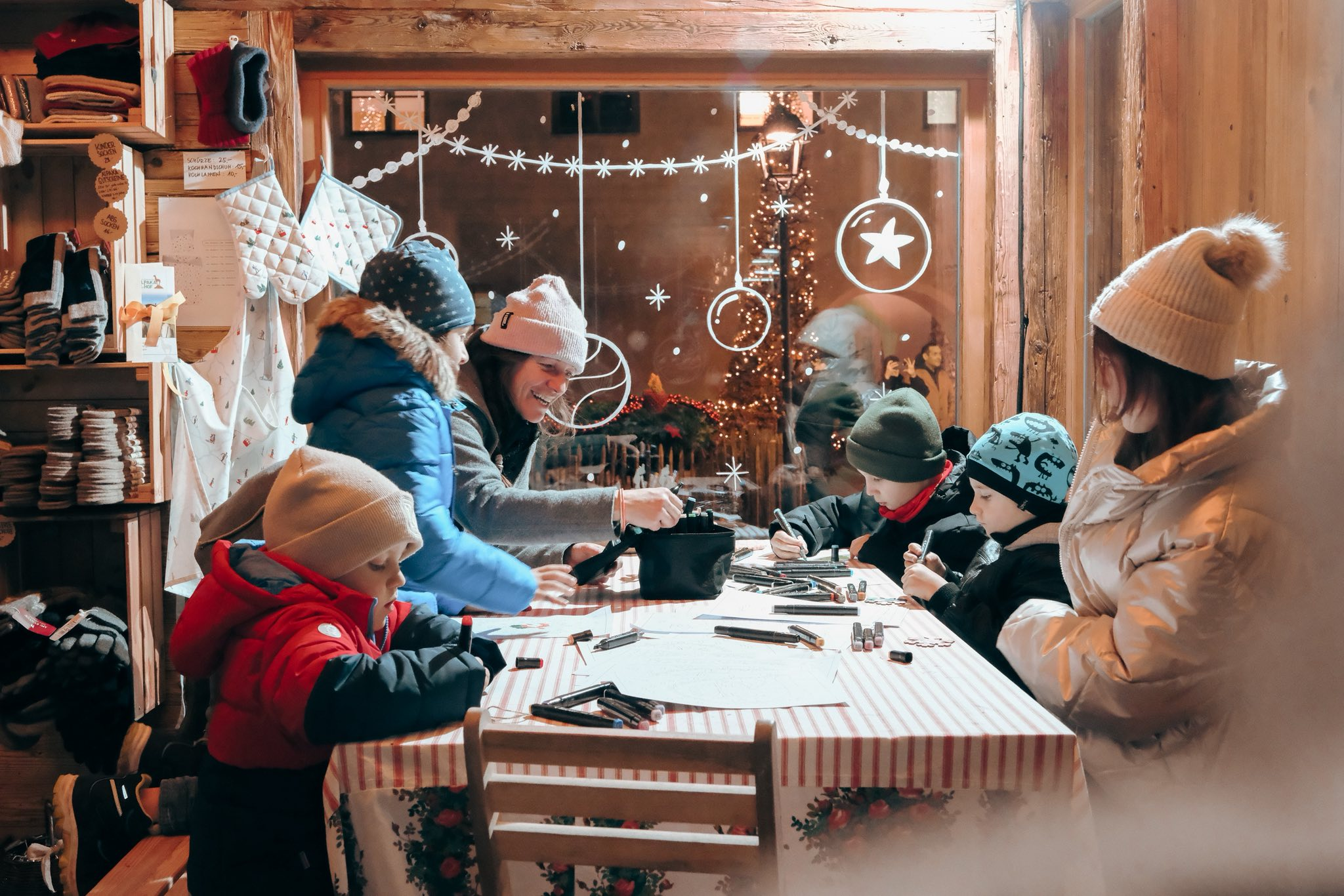 Kinderen en volwassenen in winterkleding zitten samen aan een tafel te tekenen en kleuren in een gezellige, feestelijk versierde houten hut.