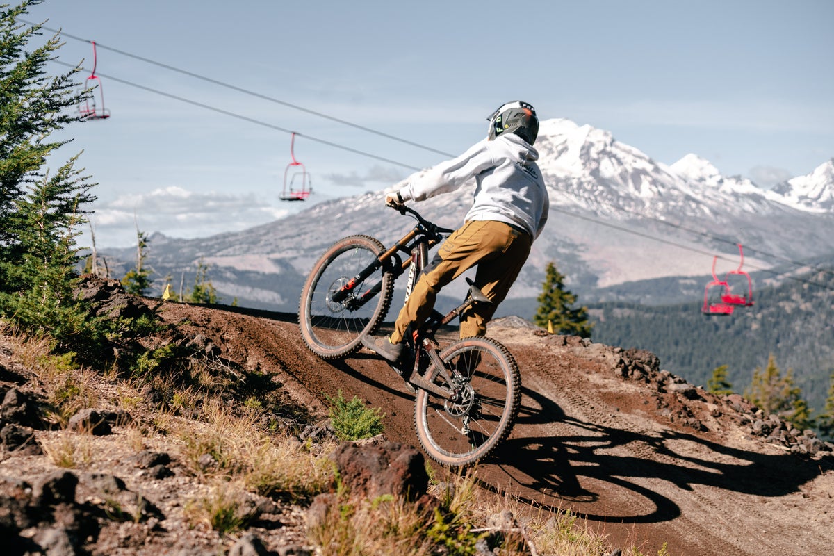 Biker riding up a hill with snow-covered mountain and chairlift in the background.