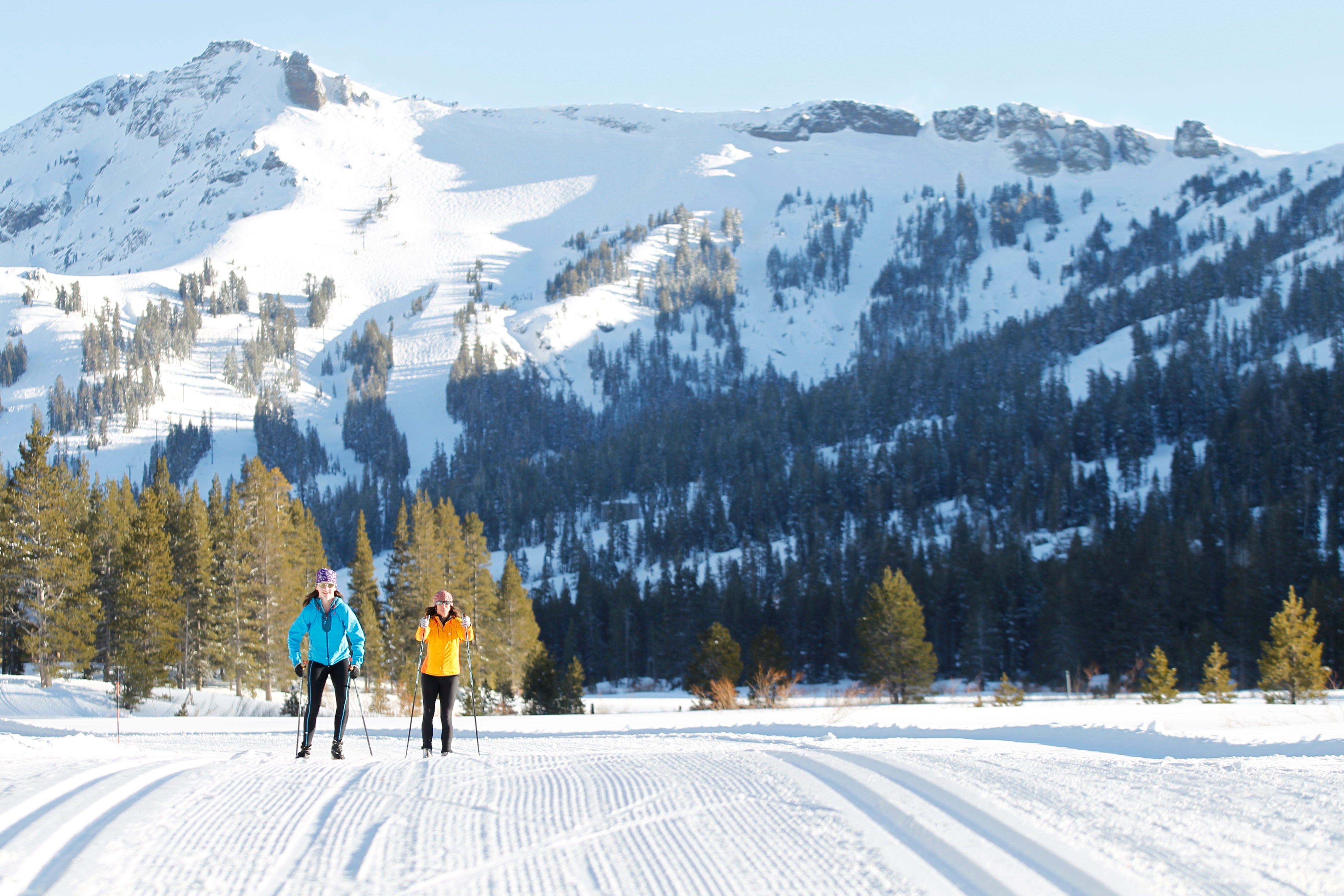 Two people skiing cross country ski trail on a sunny day with snow-covered mountain behind it