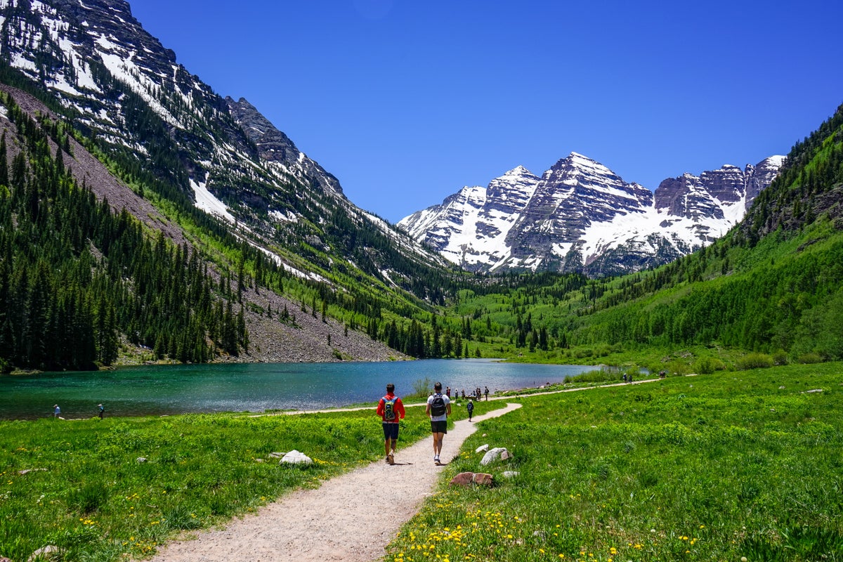 People hiking gravel trail by lake and snow-dusted mountains on a sunny day