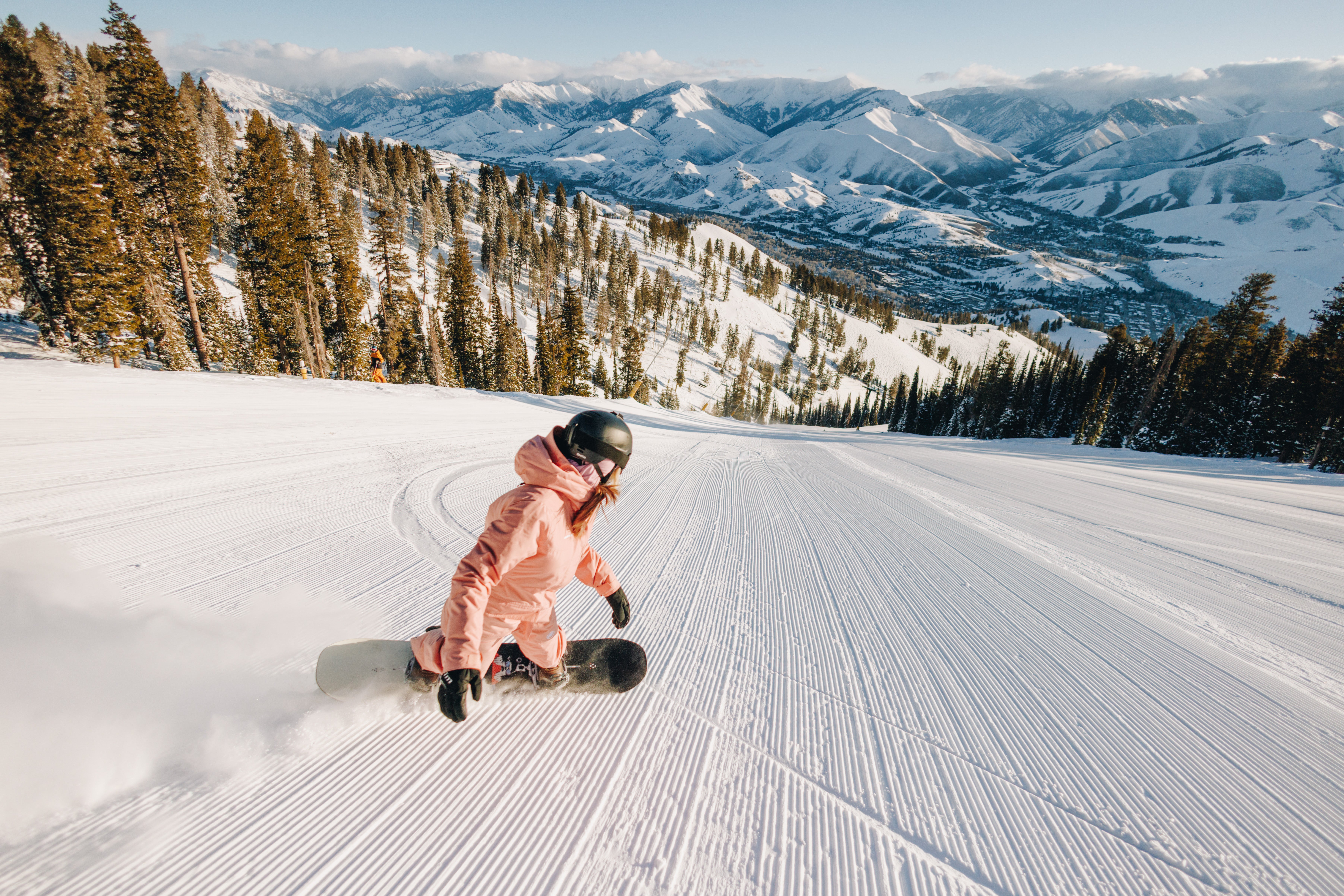 Snowboarder turning on groomed ski run with snow-covered mountains in the distance