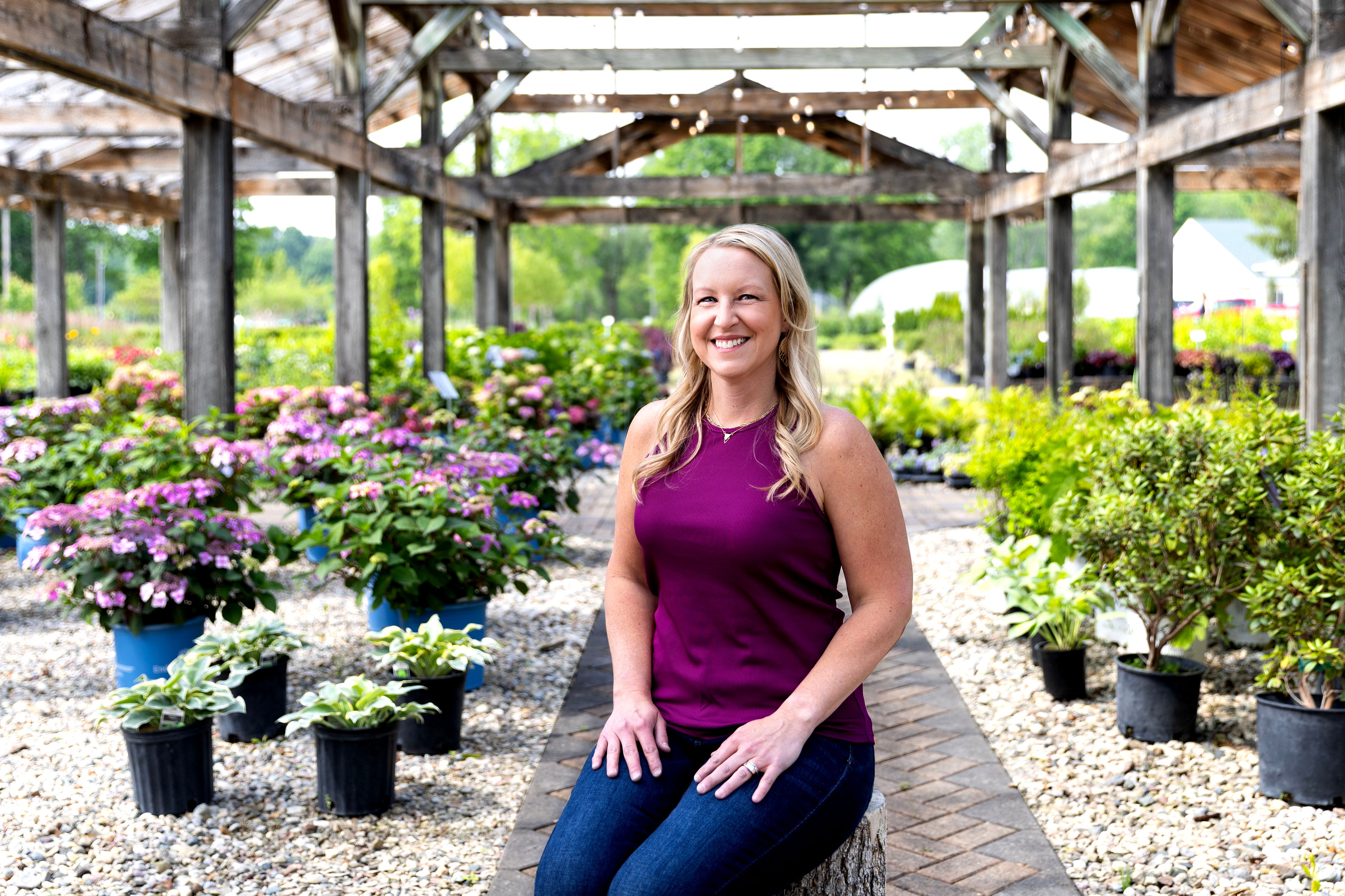 A smiling woman sitting in a greenhouse surrounded by plants.