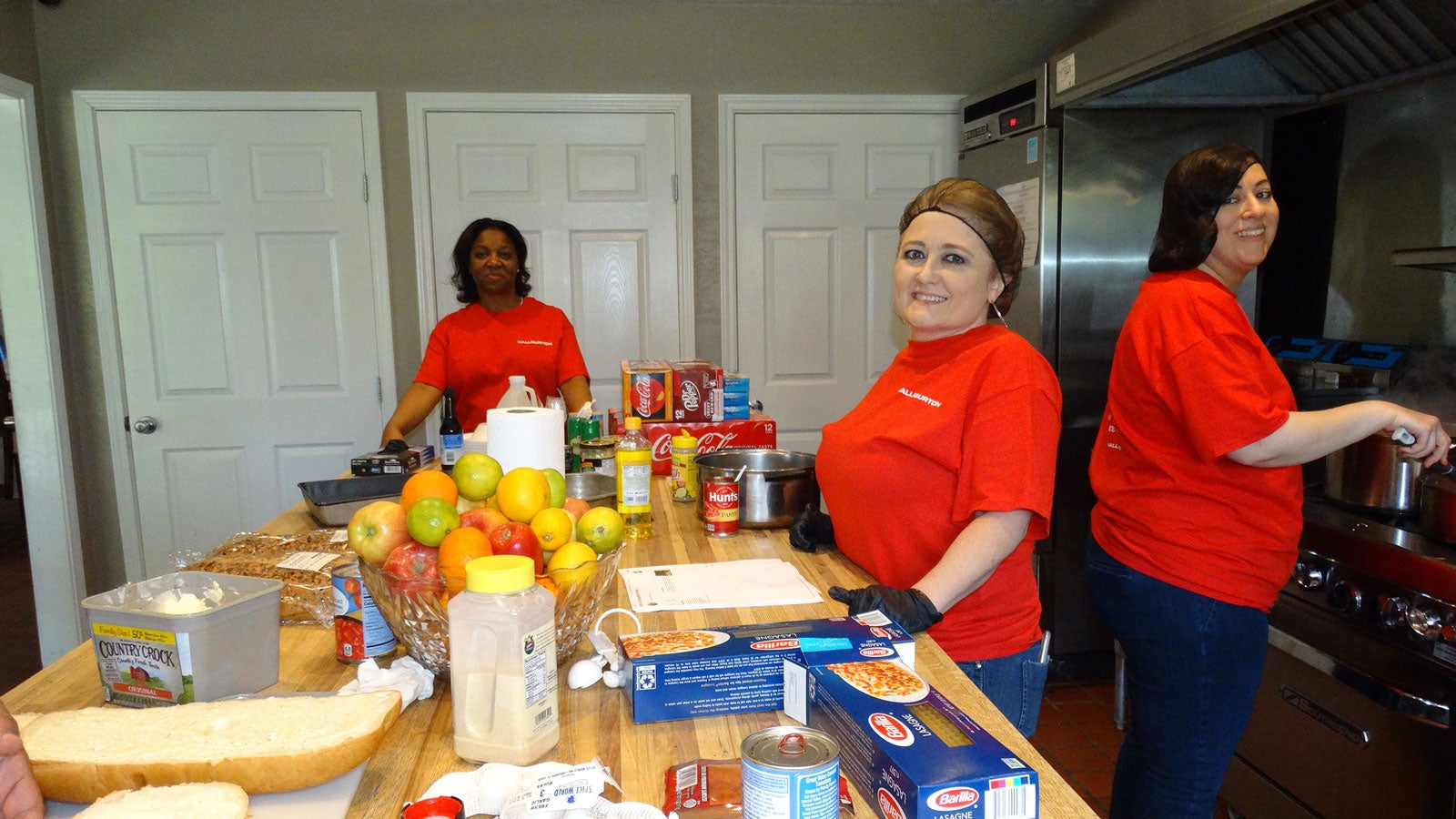 Volunteers in red shirts prepare food at a kitchen counter with bread, pasta, canned goods, and fresh produce laid out.