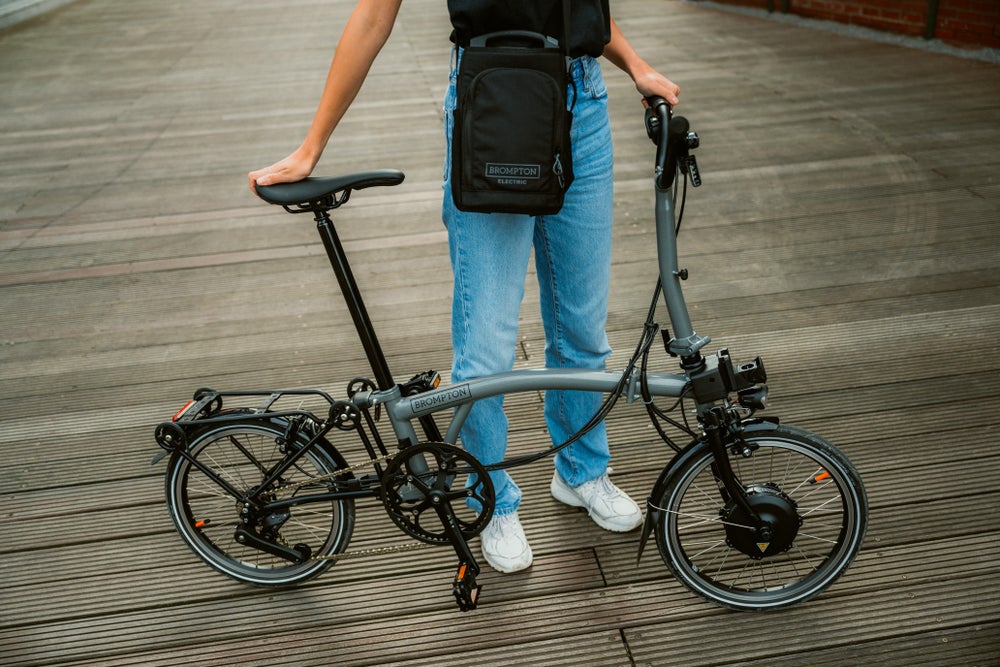 Une personne portant un vélo Brompton Electric P Line plié de couleur Storm Gray