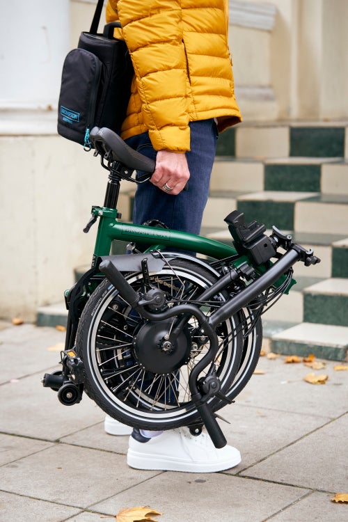 person in yellow jacket carrying folded brompton electric in forest green