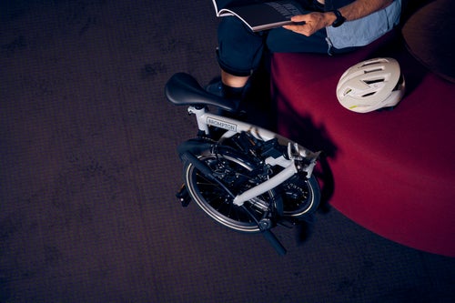 a lunar ice brompton electric p line bike folded up next to a red couch