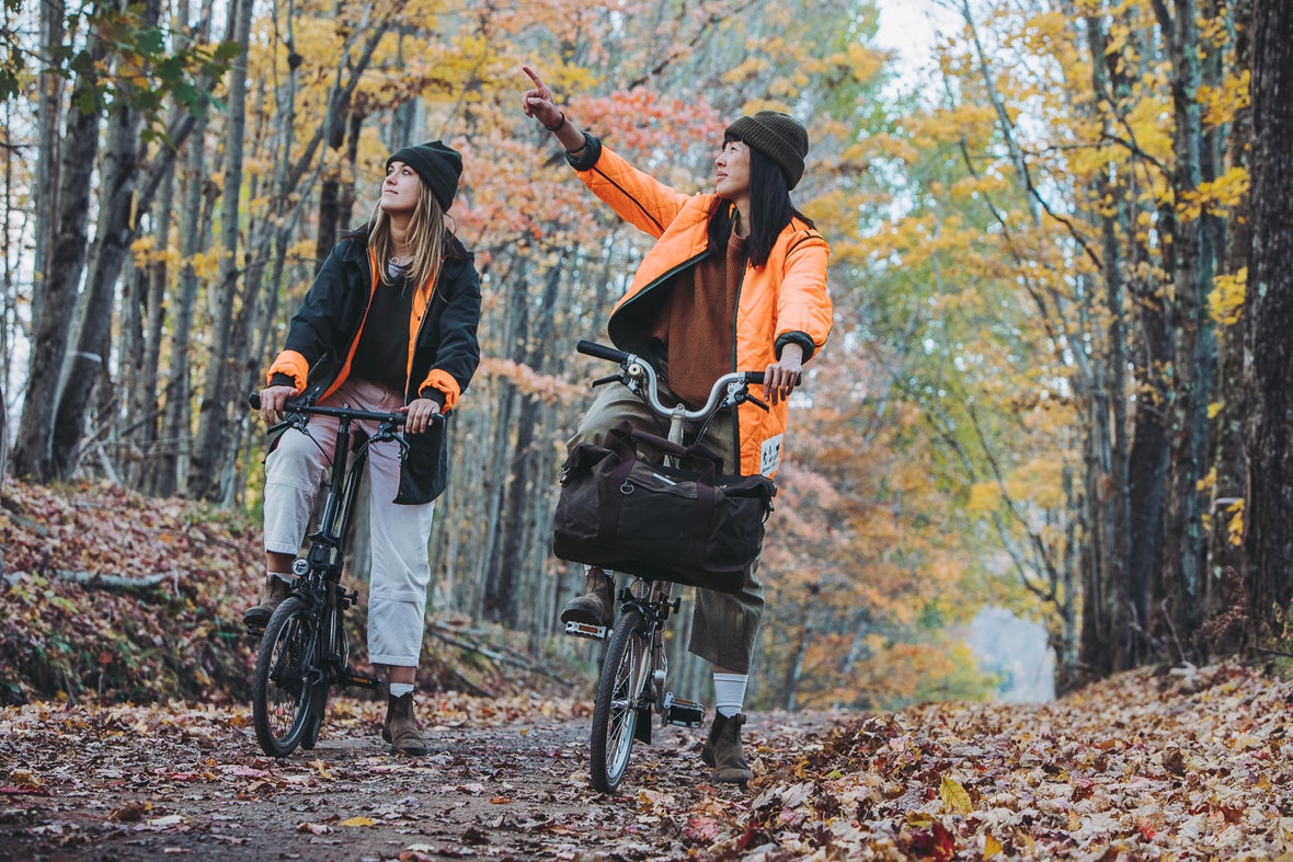 Two women on Barbour x Brompton C Lines riding through the forest in Barbour x Brompton jackets