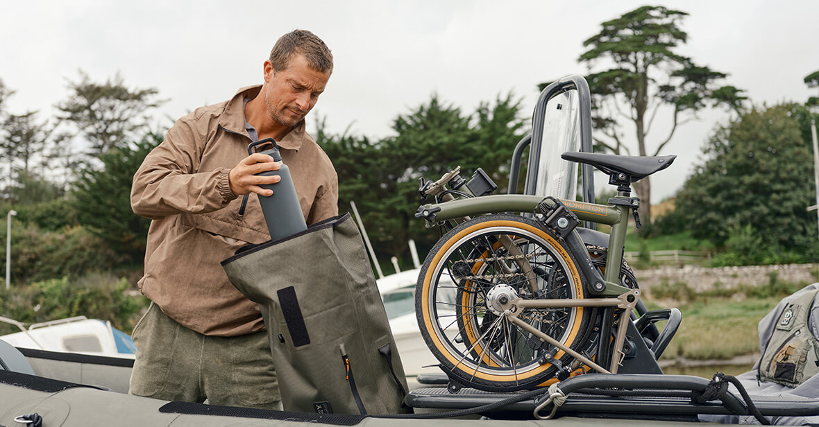 A photograph of Bear Grylls unpacking his Brompton x Bear Grylls luggage next to his folded Brompton x Bear Grylls bike
