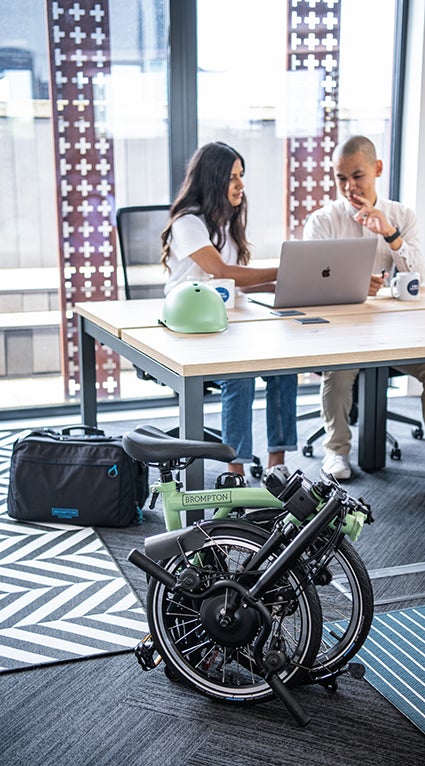 Two people working at a desk with a Brompton folded in the foreground.