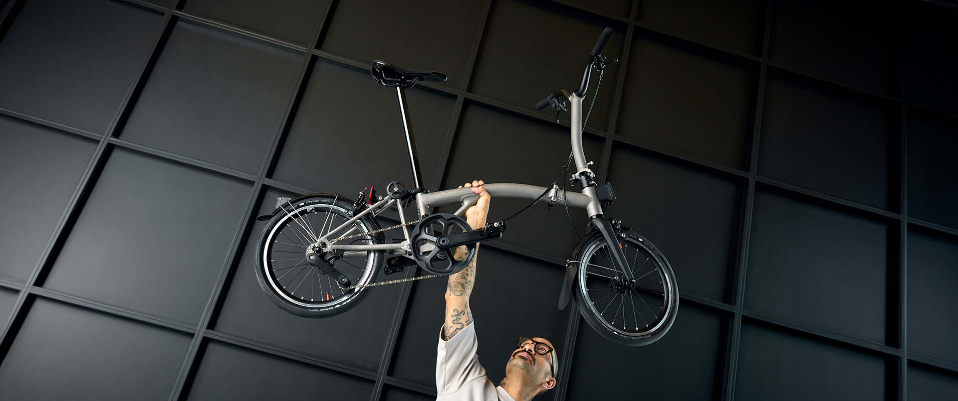 A tattooed man wearing glasses holding a Brompton T Line folding bike above his head