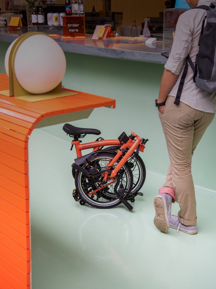 A folded Fire Coral Brompton bike next to an orange table and against a mint green wall