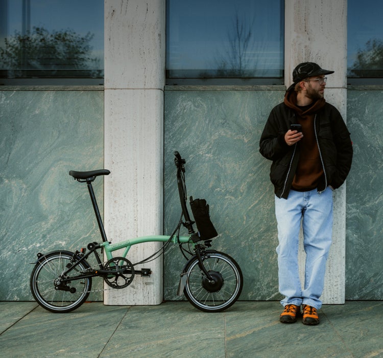 A man leaning against a wall looking at his phone next to his Brompton Electric C Line