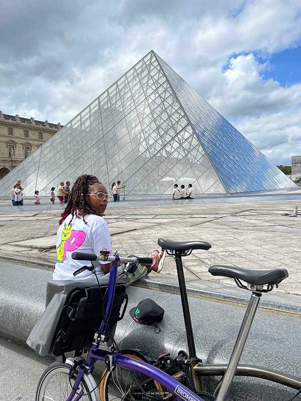 Woman sat with two Brompton bikes with the Louvre in the background