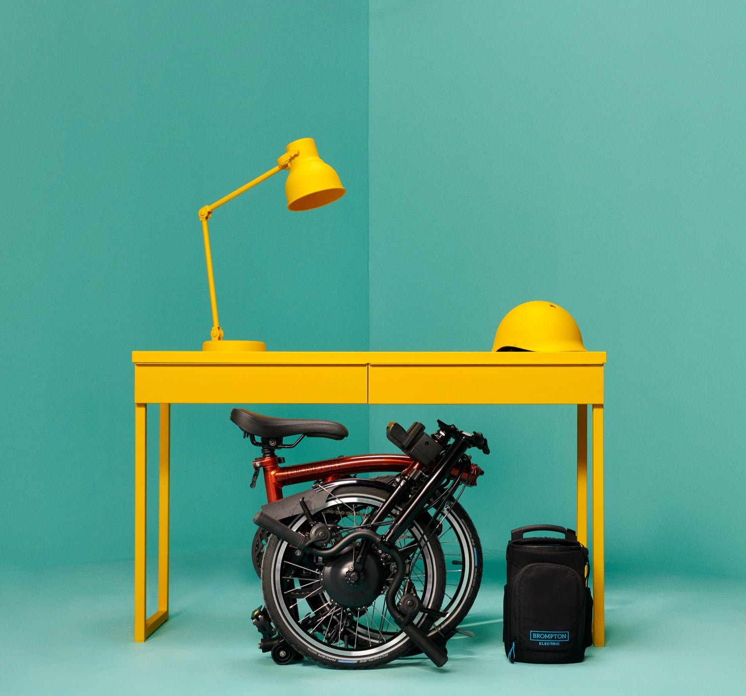 A studio shot of a folded Brompton Electric under a yellow table in a teal backdrop