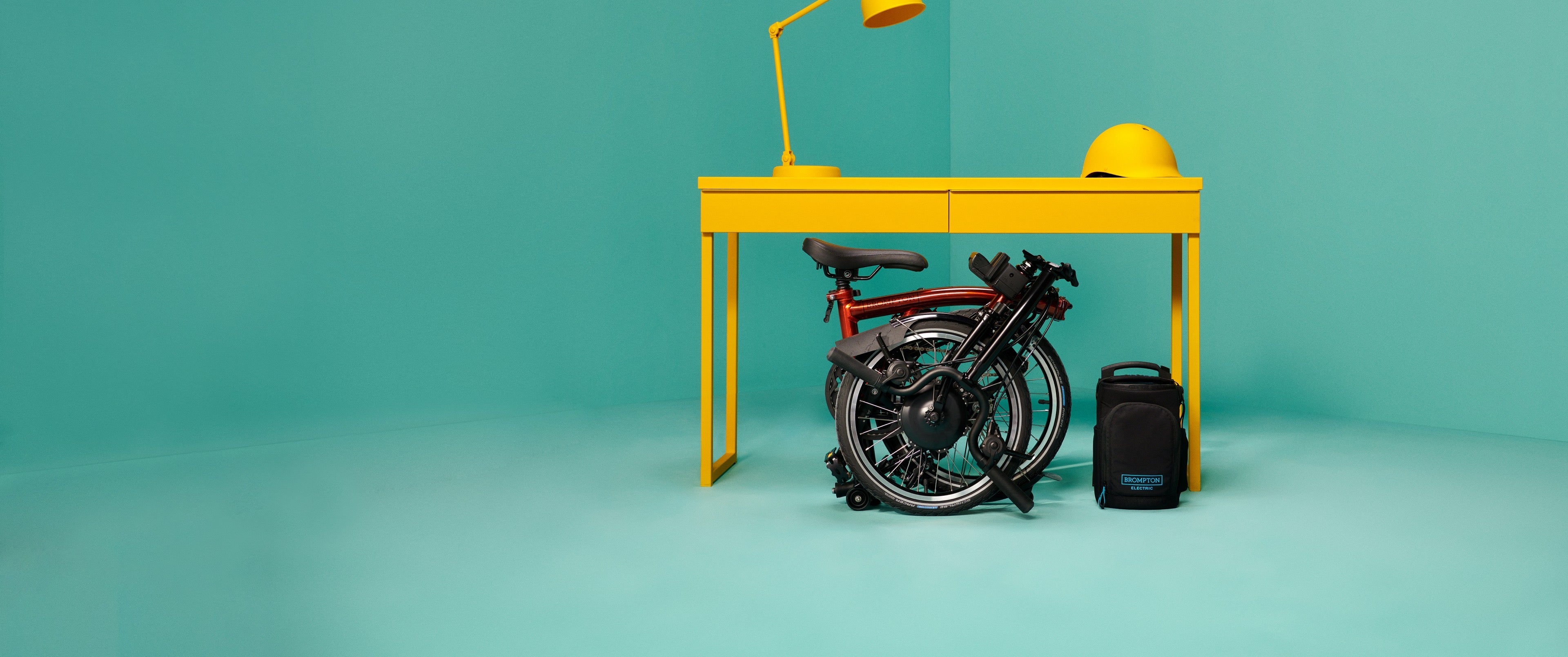 A studio shot of a folded Brompton Electric under a yellow table in a teal backdrop