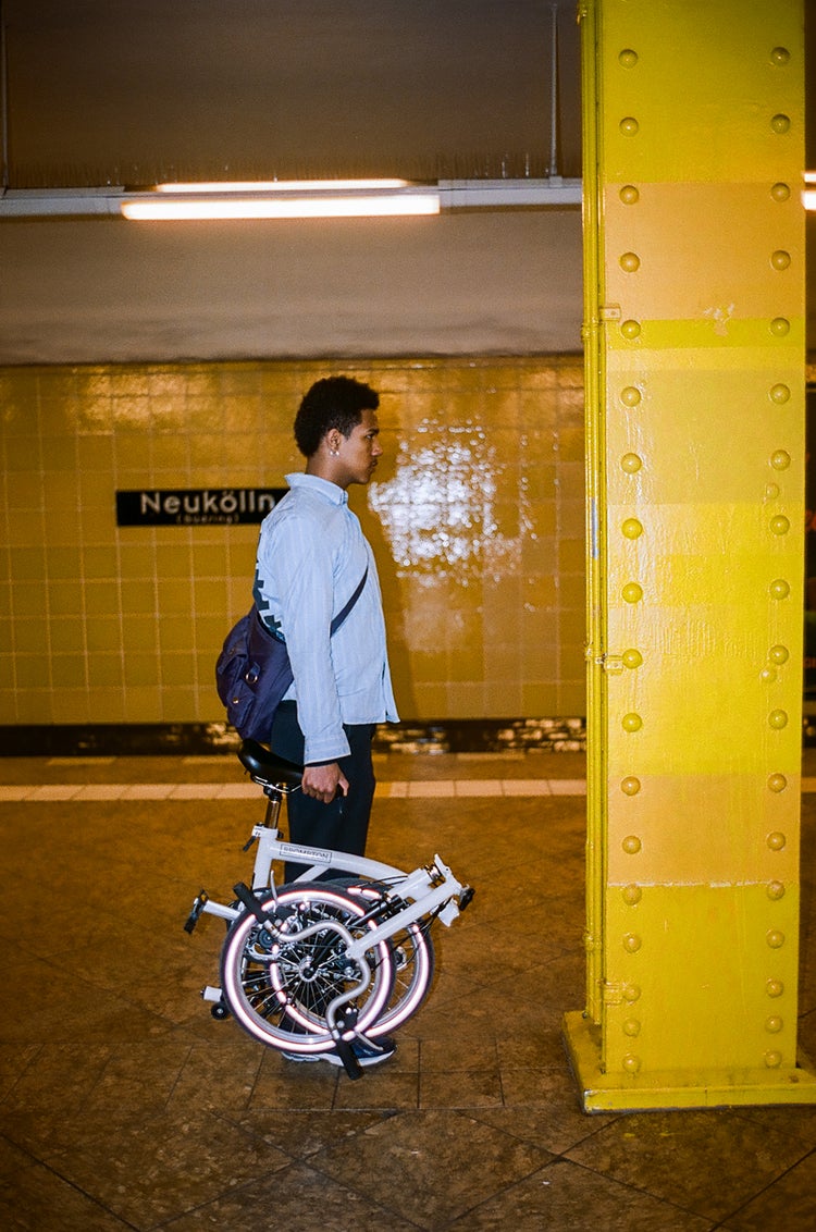 Person, die ein gefaltetes Brompton-A-Line-Fahrrad in einem gelb gekachelten Bahnhof trägt