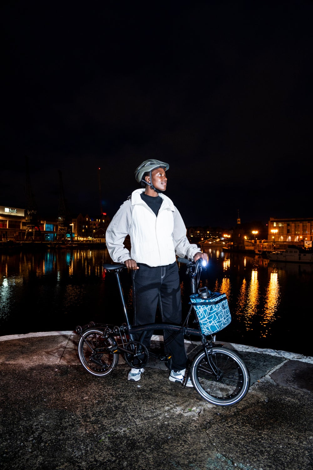 A happy person with their Brompton folding bike at night with the Bright Night luggage collection attached to the front of their bike