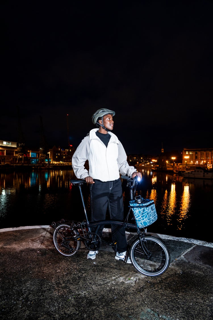A happy person riding their Brompton folding bike at night with the Bright Night luggage collection attached to the front of their bike