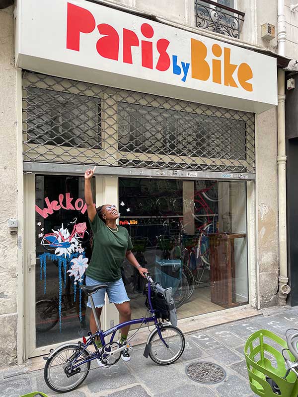 Woman stood with Brompton bike under 'Paris by Bike' sign