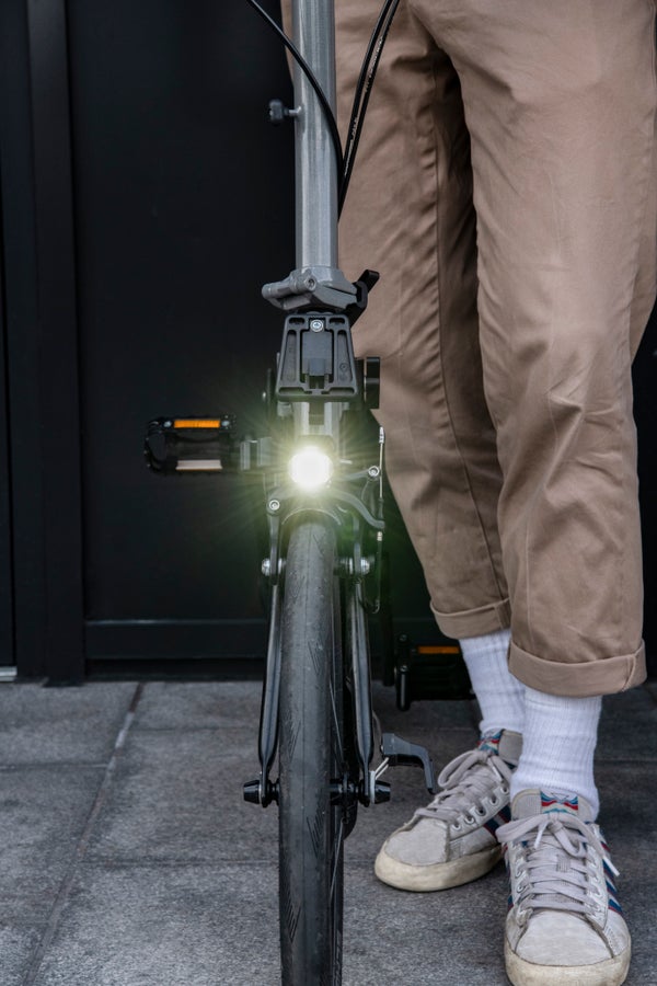 A photograph of a person standing next to their Brompton bike with the 500lm front light shining bright