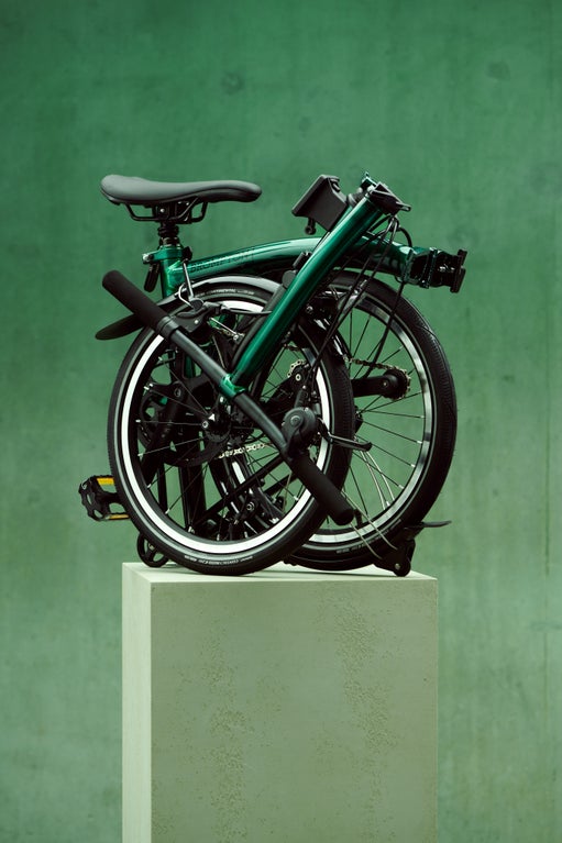 A smiling woman carrying a Brompton Electric C Line in a teal studio background