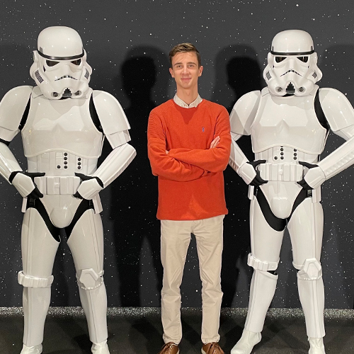 Disney UK Intern, Oliver, wearing khaki pants and an orange shirt, smiling while standing between two Star Wars Storm Troopers