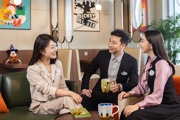 three young Asian people in business attire, seated around a coffee table in a room decorated with Disney memorabilia
