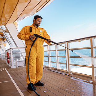 male crew member in an orange jumpsuit uniform, powerwashing the deck